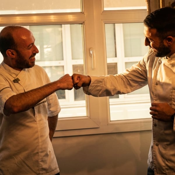 Two smiling chefs in white jackets fist-bumping near a sunlit window.