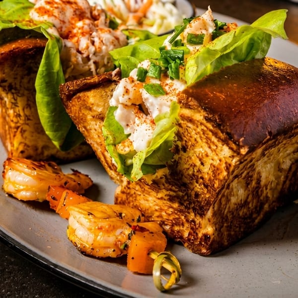 Two lobster rolls on brioche bread with lettuce, shrimp skewer, and coleslaw on a gray plate.
