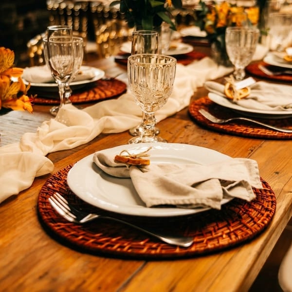 Elegantly set dining table with white plates, woven placemats, crystal glasses, and orange floral centerpieces.