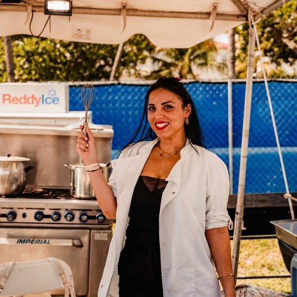 A smiling chef in a white coat holds a whisk at an outdoor cooking station with a ReddyIce sign.